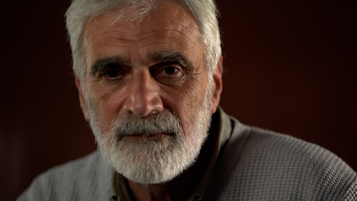 A grey-bearded man in a grey sweater against a deep red wall (1/3)