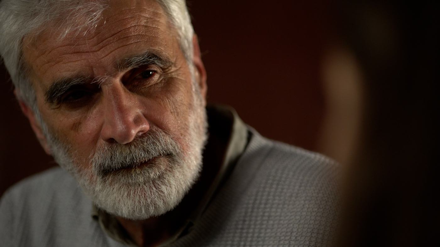 A grey-bearded man in a grey sweater against a deep red wall (2/3)