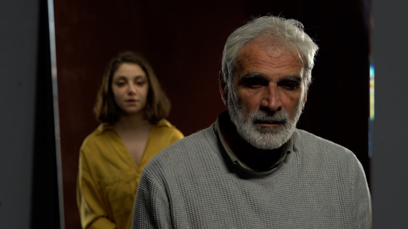 A grey-bearded man in a grey sweater against a deep red wall (3/3)